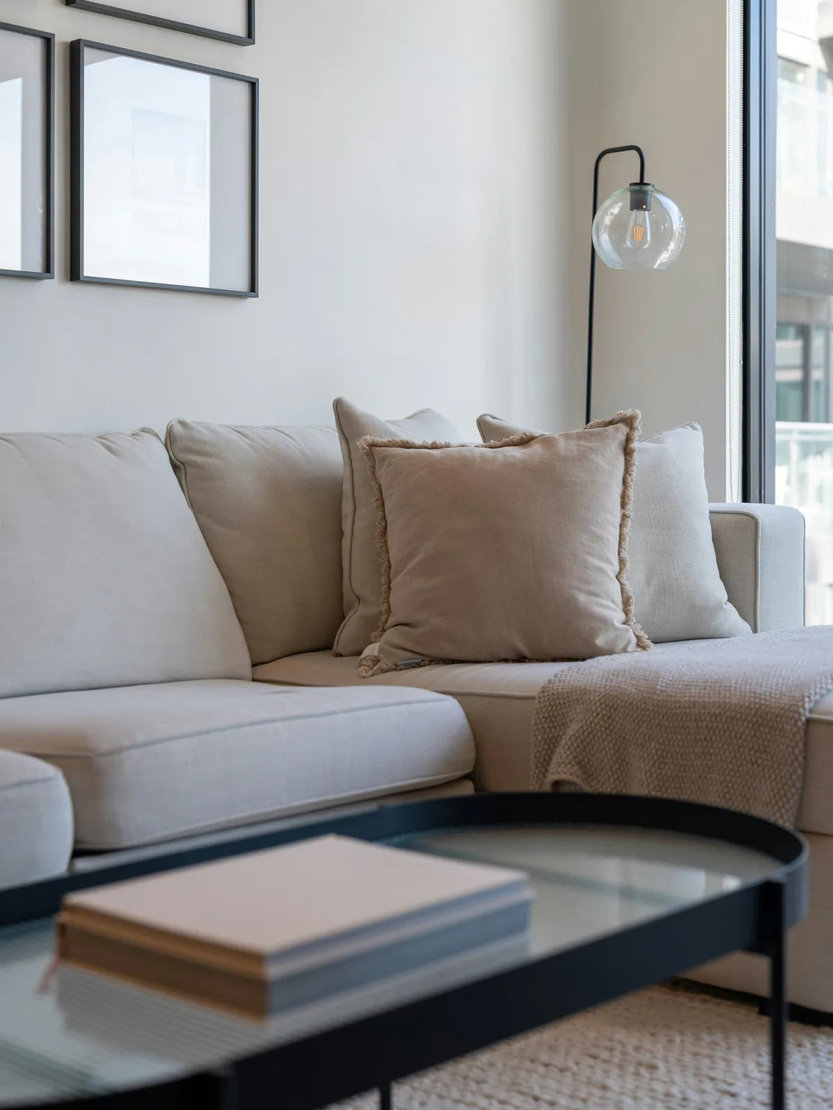 A pale sectional sofa with linen cushions, a black globe floor lamp beside it, and a glass-topped oval table in front of a full-height window.